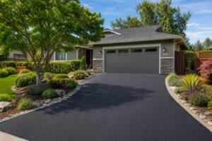 Wide, landscape view of a newly paved residential asphalt driveway in a Bay Area suburban home, featuring smooth blacktop, landscaped garden beds, and a modern house with a two-car garage.