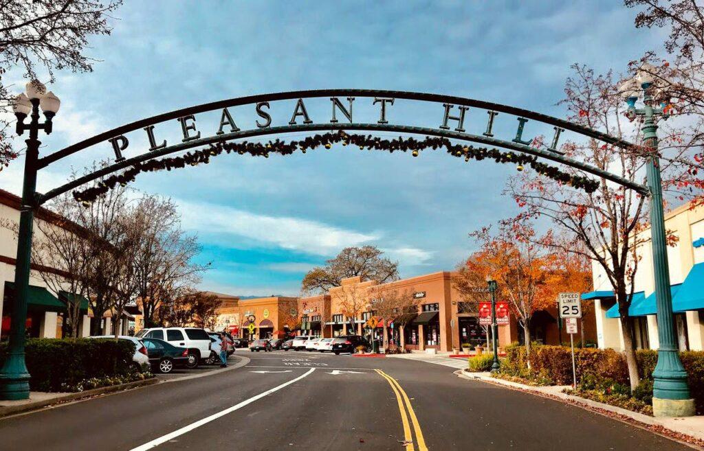 Pleasant Hill downtown street scene featuring decorative archway, local shops, parked cars, and tree-lined sidewalks under a cloudy sky.
