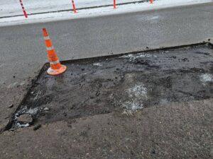 Damaged asphalt roadway section marked with a traffic cone during winter road repair work, showing deteriorated pavement, surface wear, and patch preparation in cold weather conditions.