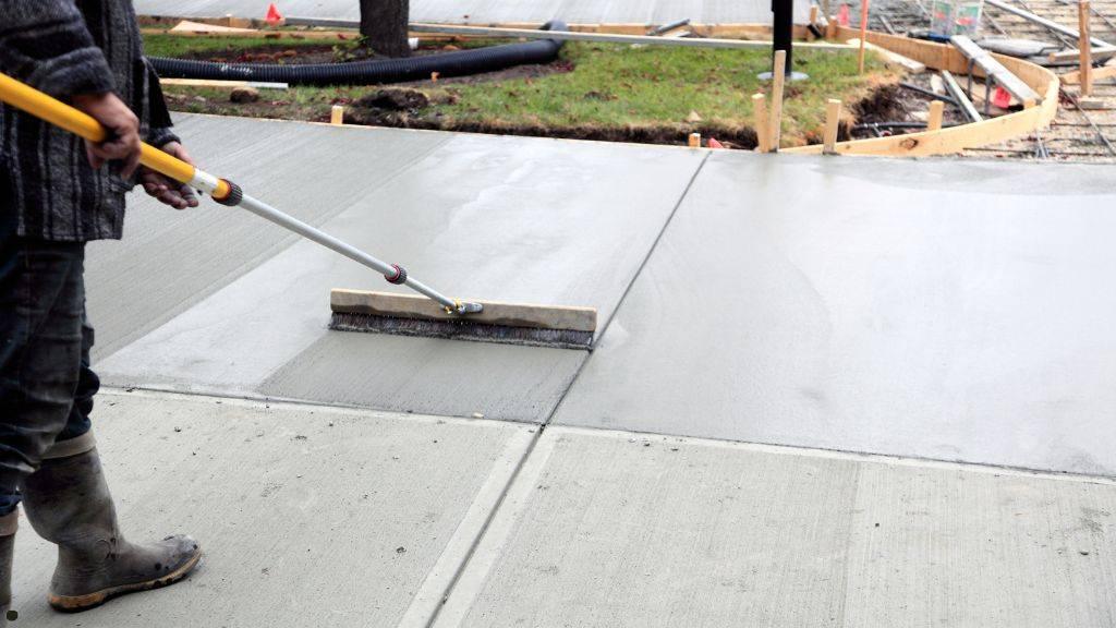 Construction worker using a concrete broom to apply a slip-resistant texture to a newly poured walkway.
