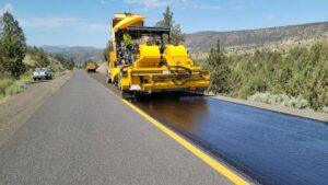 Yellow asphalt chip spreader machine applying fresh chip seal surface treatment along a rural private road before winter.
